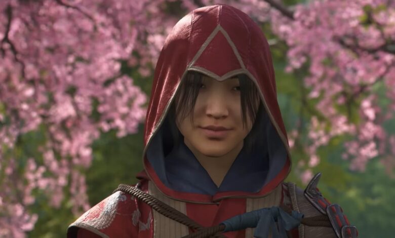 Close-up of a young woman in a red hooded Assassin's Creed outfit, set against a blurred background of pink cherry blossoms.