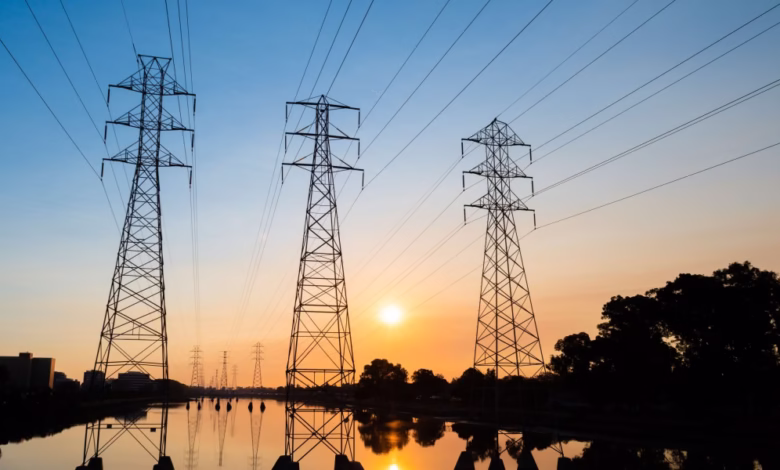 Three tall electricity pylons stand silhouetted against a vibrant sunset over calm water.