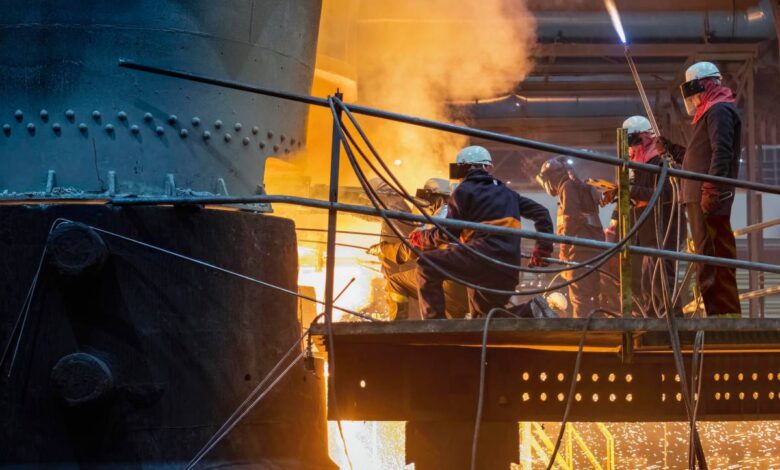Workers in protective gear use torches near a glowing metal furnace, sparks flying.