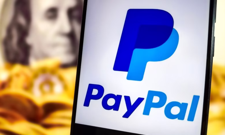 Close-up of a smartphone displaying the PayPal logo, with a blurred image of a US $100 bill in the background.