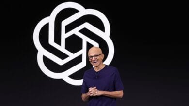 Satya Nadella, Microsoft CEO, smiles on stage before a large white logo on a black background.