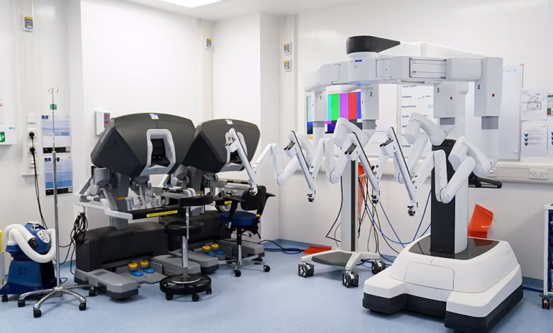 Two robotic surgical systems in a modern operating room. One is a large, white robotic arm; the other is a dark gray console with two robotic arms.