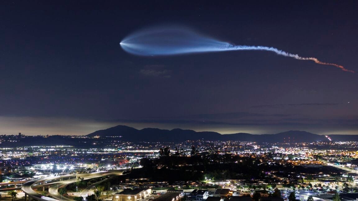 A SpaceX Falcon 9 rocket's trail illuminates the night sky above a city skyline.