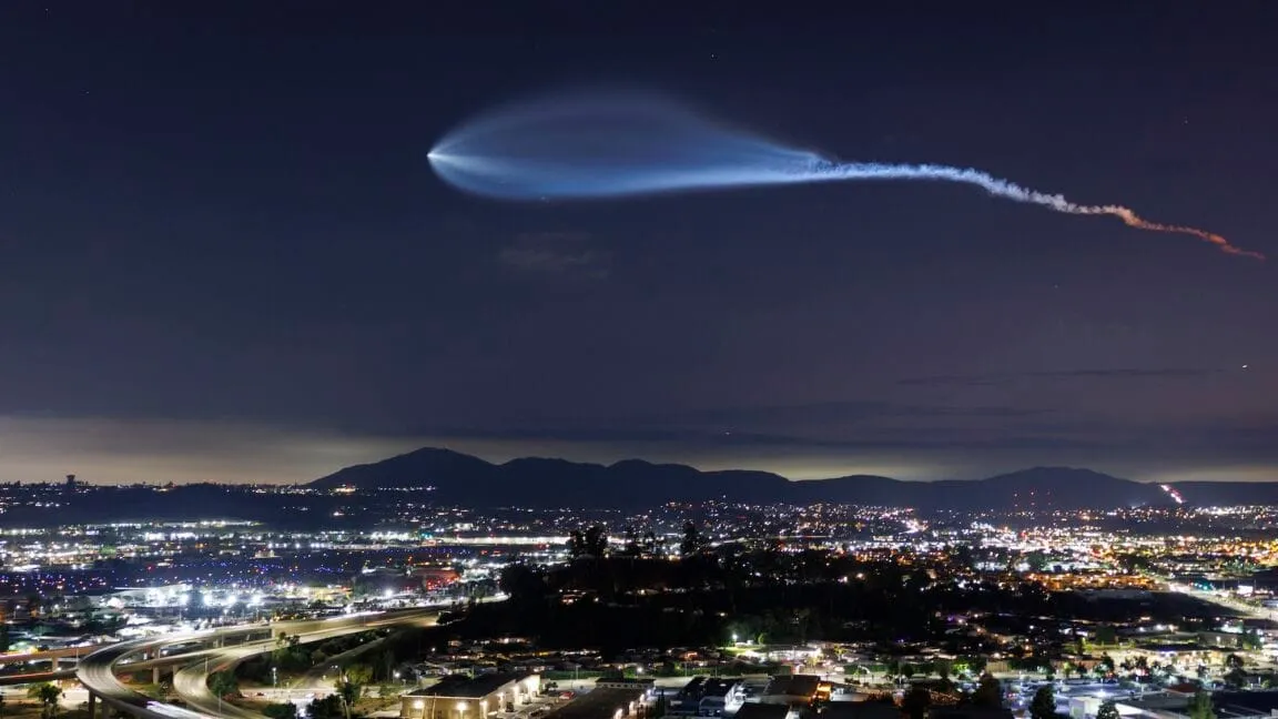 A SpaceX Falcon 9 rocket's trail illuminates the night sky above a city skyline.