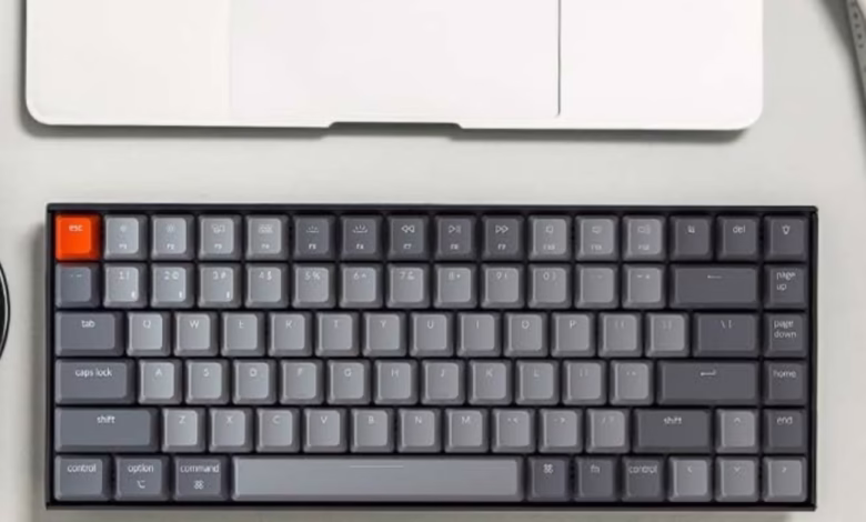 Compact dark gray Keychron K2 keyboard with an orange escape key sits on a desk next to a laptop and phone.
