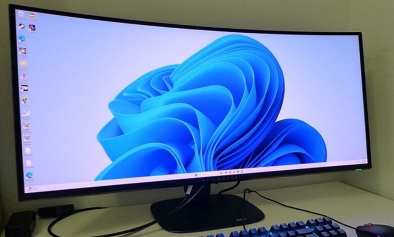 Curved computer monitor displaying the Windows 11 default background, a vibrant blue abstract design. A keyboard and mouse are partially visible.