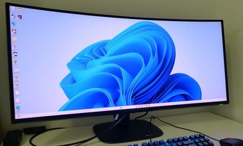 Curved computer monitor displaying the Windows 11 default background, a vibrant blue abstract design. A keyboard and mouse are partially visible.
