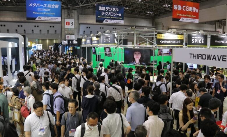A large crowd of people at a technology expo, many wearing masks, milling around booths with screens displaying digital content.