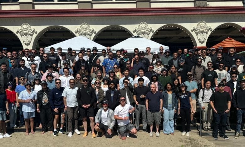 Large group photo of the Observe team on a sunny beach, many wearing matching bucket hats.