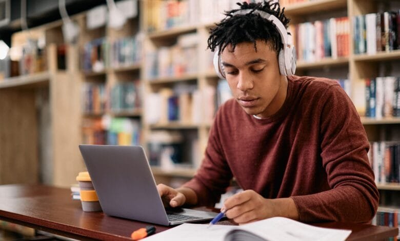 Focused young man studies at a library table, using a laptop and taking notes. Bookshelves blur in the background.