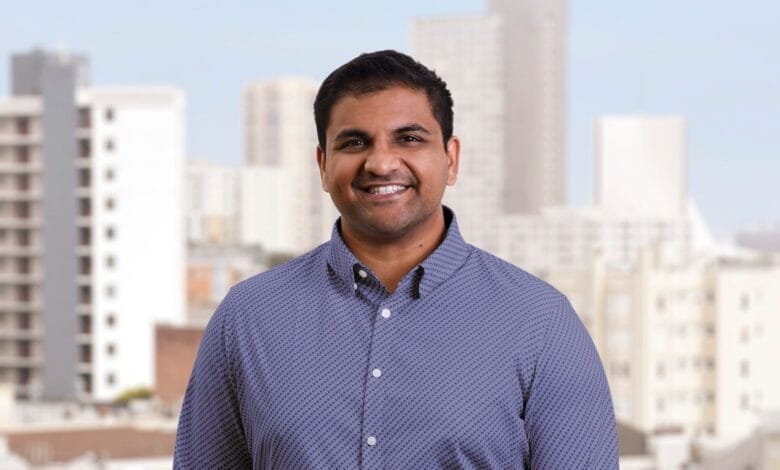 Smiling man in a blue patterned shirt stands before a blurred cityscape.