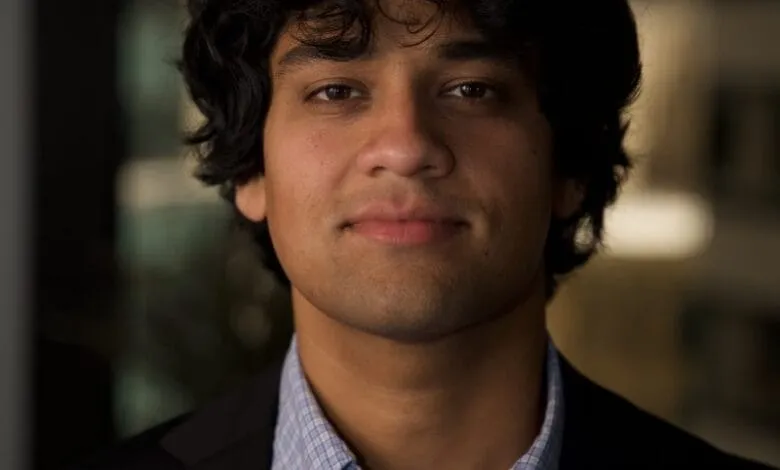 Headshot of a young man with dark curly hair, wearing a dark suit jacket over a light blue plaid shirt.