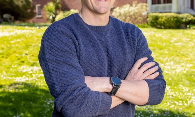 Man with arms crossed smiles outdoors, wearing a blue sweater and gray pants. A brick building is in the background.