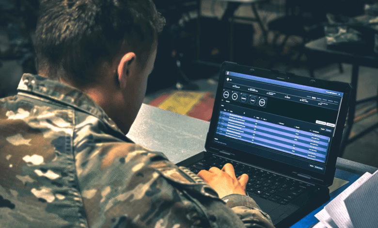 A soldier in camouflage reviews data on a laptop with a dark interface, possibly related to military operations or logistics.
