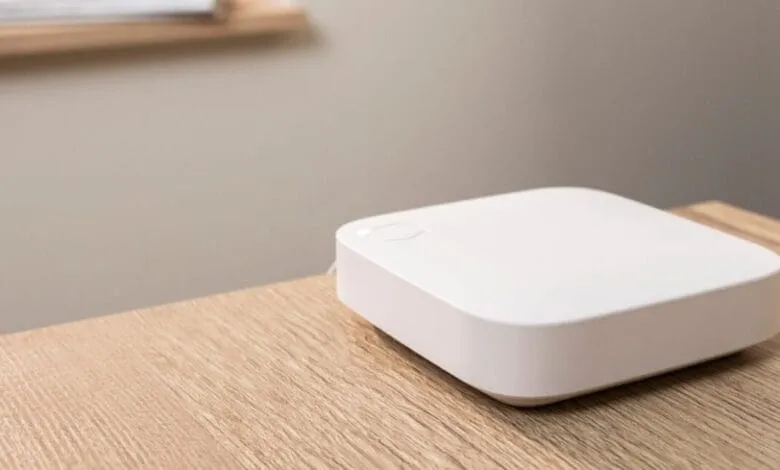 A small, white, square device sits on a light wood table. It appears to be a smart home hub.