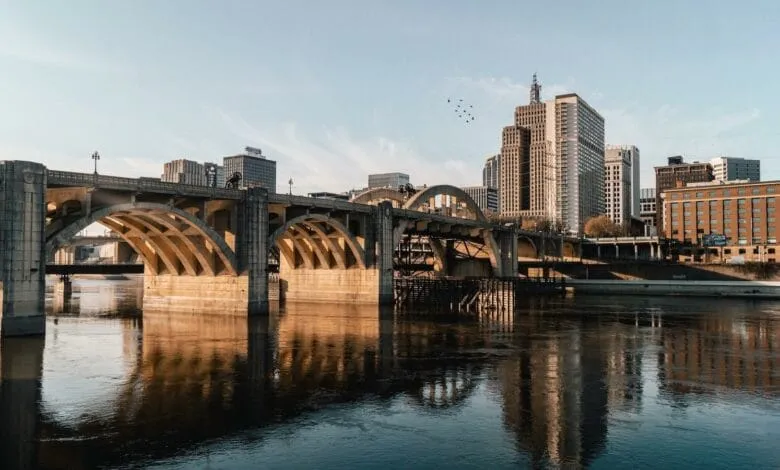 A long, arched bridge spans a river, reflecting the city skyline of St. Paul, Minnesota, under a clear sky.