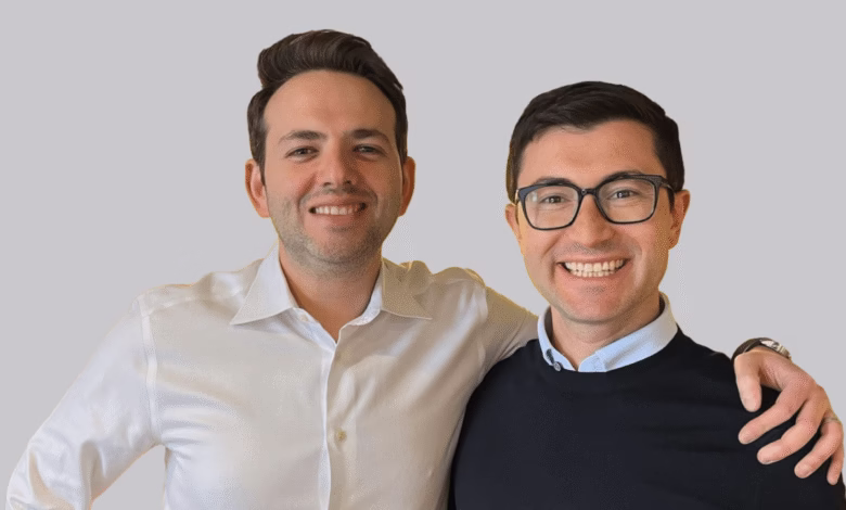 Two smiling men stand shoulder to shoulder, one in a white shirt and the other in a dark sweater.