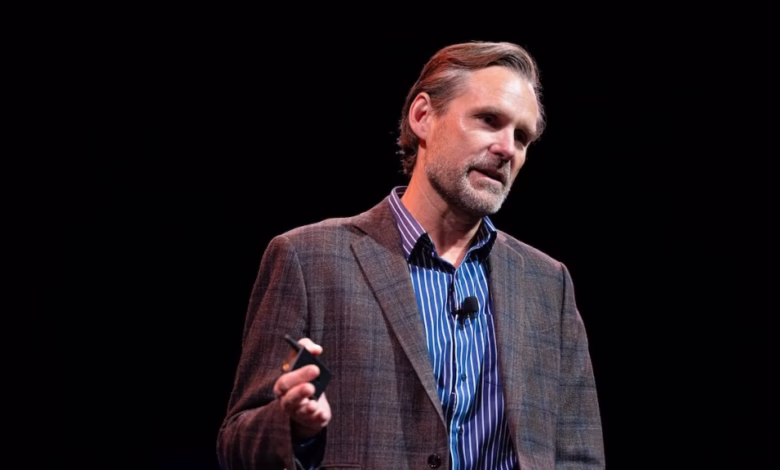 A man in a brown blazer and blue striped shirt speaks at a conference, holding a small black device.