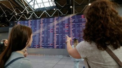 Two women check a large airport departures board showing numerous flight delays or cancellations.