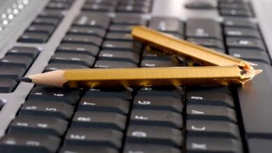 Two broken gold pencils lie on a black computer keyboard, symbolizing writer's block or technological frustration.