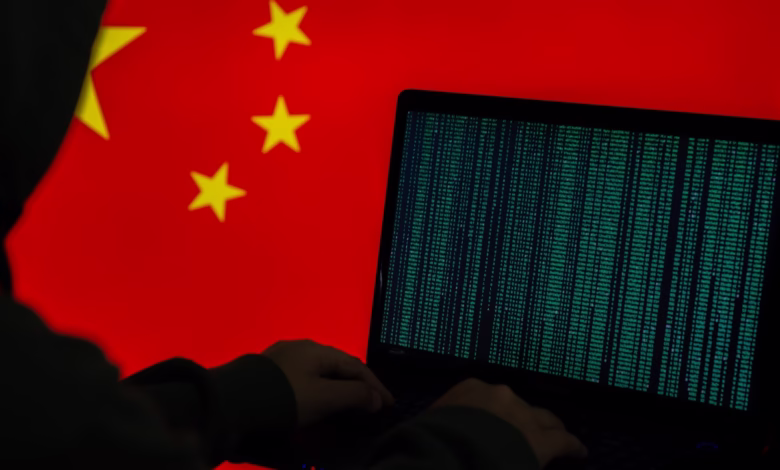 A hooded figure sits before a laptop displaying green code, with a blurred Chinese flag in the background, suggesting cyber activity.
