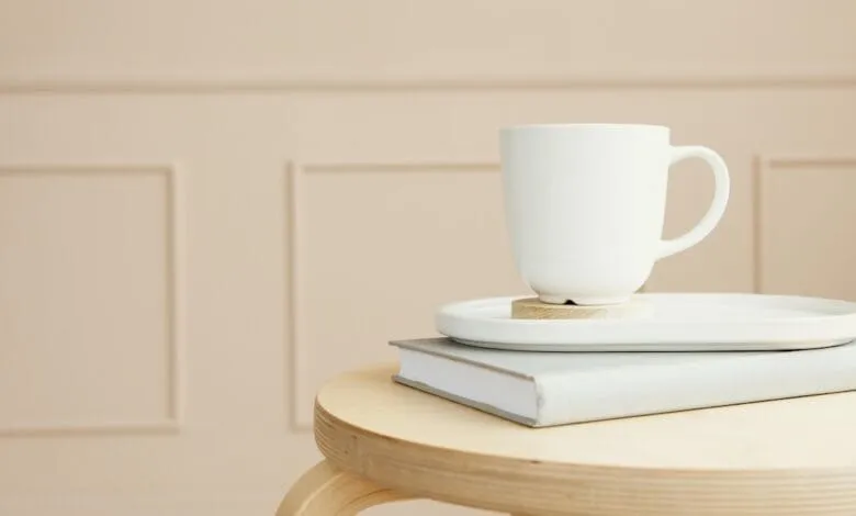 A white mug rests on a gray coaster atop a book on a light wood table against a beige wall.