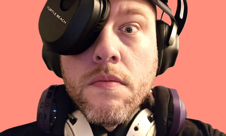 Close-up of a man with a beard wearing multiple headsets, one covering his left eye. The coral background contrasts with the dark headsets.