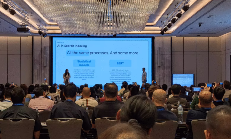 A large conference room with a presentation on AI in search indexing. Two speakers present to a large audience.