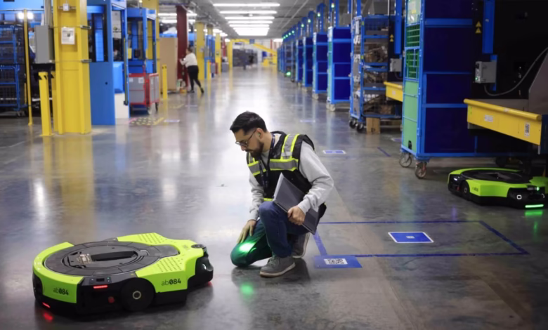 A warehouse worker kneels beside a small, bright green robot, inspecting it on a polished concrete floor. Other robots and industrial equipment are visible in the background.