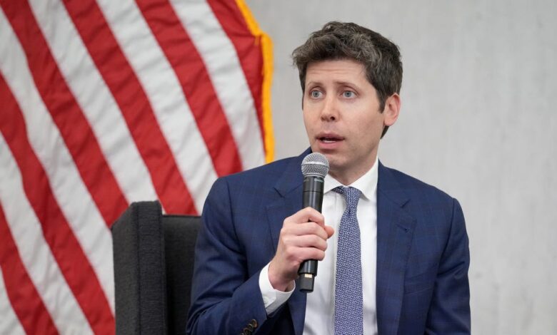 A man in a blue suit speaks into a microphone, with an American flag in the background.