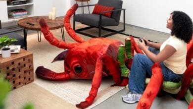 A young woman plays video games while sitting on a giant, plush ant. The ant is red and green, and the room is modern and minimalist.