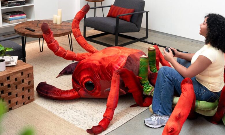 A young woman plays video games while sitting on a giant, plush ant. The ant is red and green, and the room is modern and minimalist.