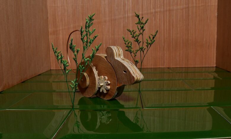 Wooden toy mouse with gears, positioned on dark green tiles near small green plants against a wood backdrop.