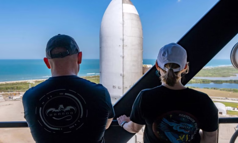 Two people wearing black t-shirts with space-themed logos stand on a balcony overlooking a large rocket and ocean.