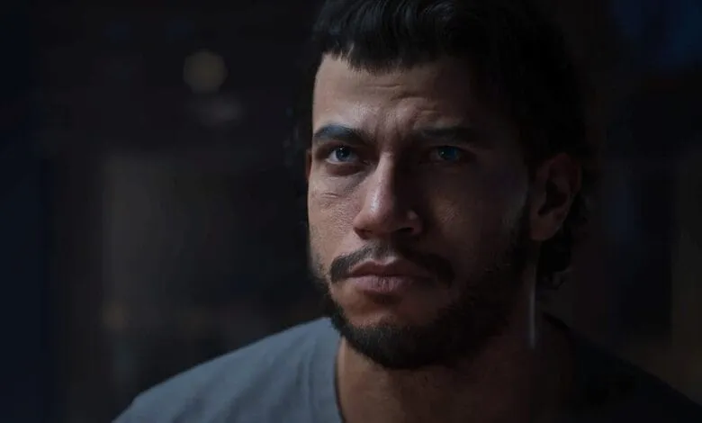 Close-up portrait of a man with intense blue eyes and a serious expression, dark hair, and a short beard.