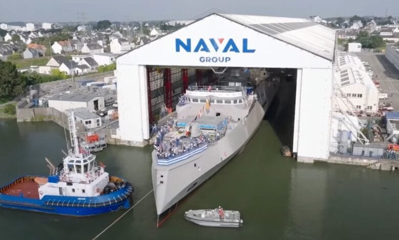 A gray warship emerges from a Naval Group shipyard, flanked by a tugboat and smaller vessel. People line the ship's deck.
