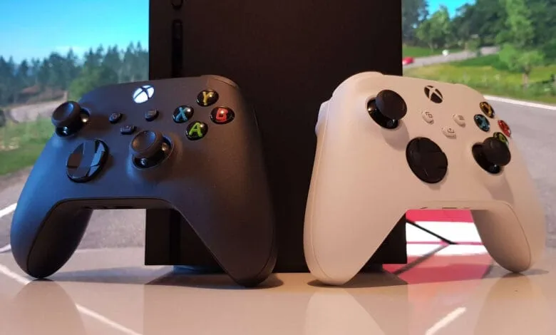 Black and white Xbox Series X controllers rest on a surface in front of the console, with a blurred racing game background.