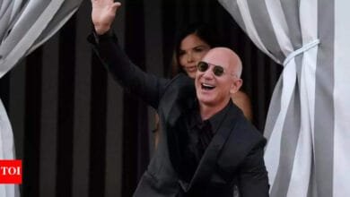Jeff Bezos, Amazon founder, waves and smiles while wearing sunglasses and a black suit.