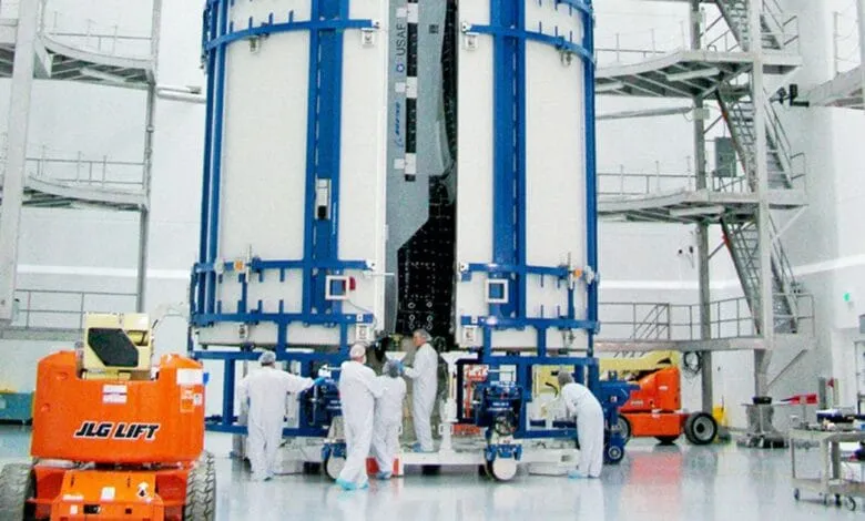 Technicians in white suits prepare a large, white spacecraft inside a hangar. Blue metal framework surrounds the craft.