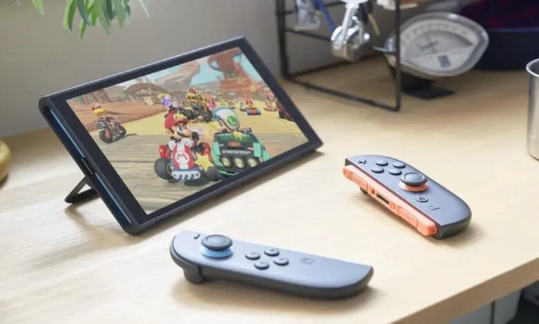 Nintendo Switch OLED screen displaying Mario Kart, with two Joy-Con controllers on a wooden desk.