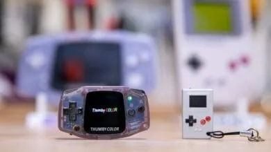 Two miniature handheld game consoles sit on a wooden surface, with blurred retro gaming devices in the background.