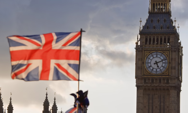 A British flag waves gently in the foreground, with the Elizabeth Tower (Big Ben) and part of the Houses of Parliament visible in the background under a cloudy sky.