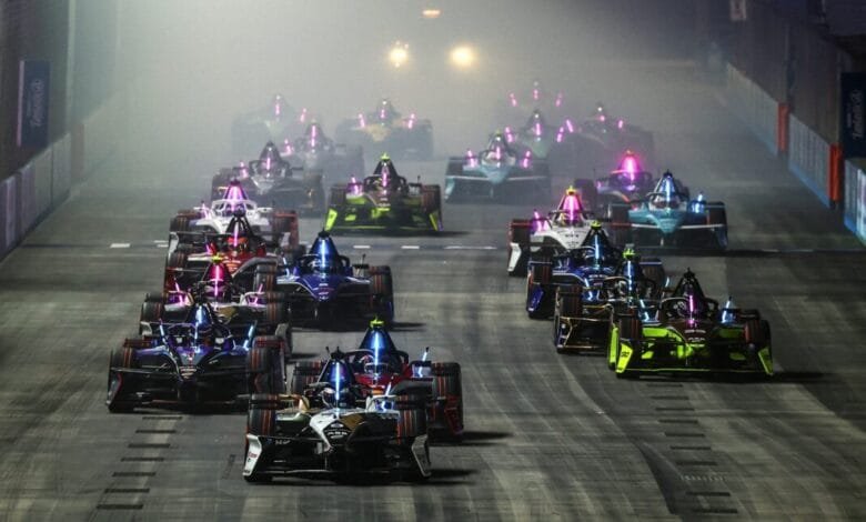 A pack of Formula E race cars speeds into the first turn, illuminated by glowing lights on their chassis. Smoke fills the track.