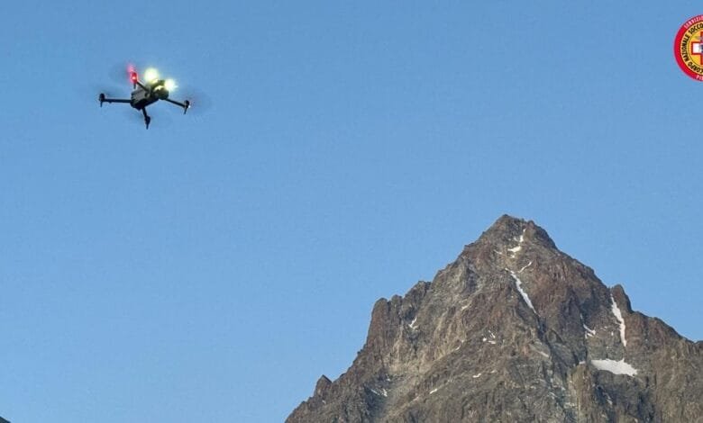 Drone flying over a mountain range under a clear blue sky. A rescue service logo is visible.