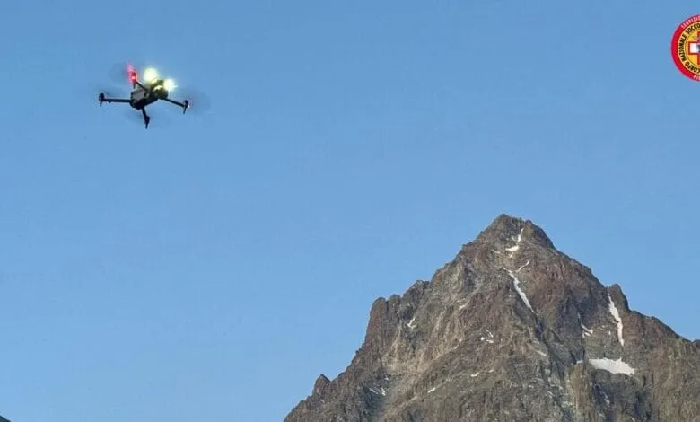 Drone flying over a mountain range under a clear blue sky. A rescue service logo is visible.