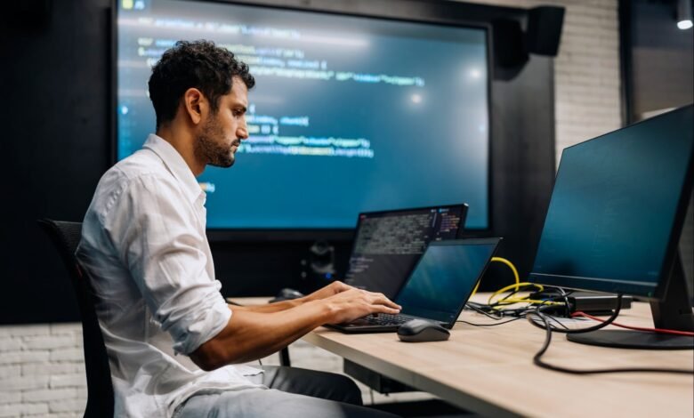 Focused programmer coding on laptop, large screens display code in the background.