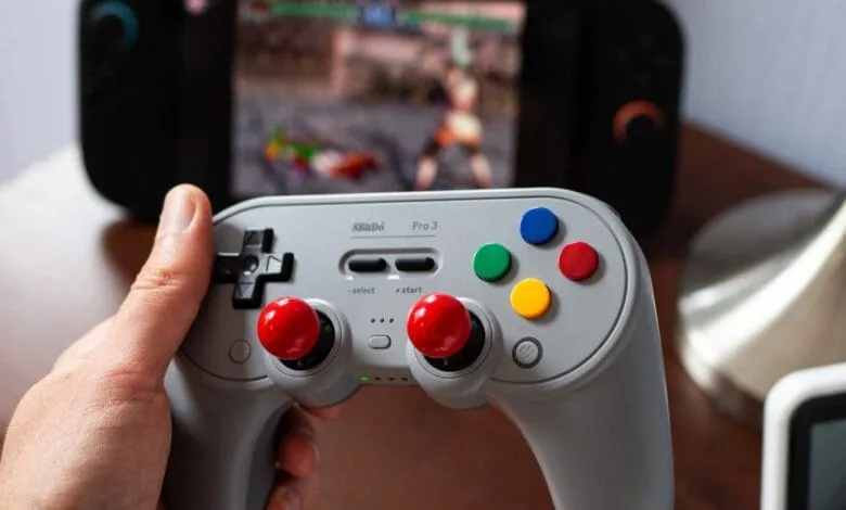 Close-up of a hand holding an 8BitDo Pro 3 controller, with a blurry video game screen in the background.