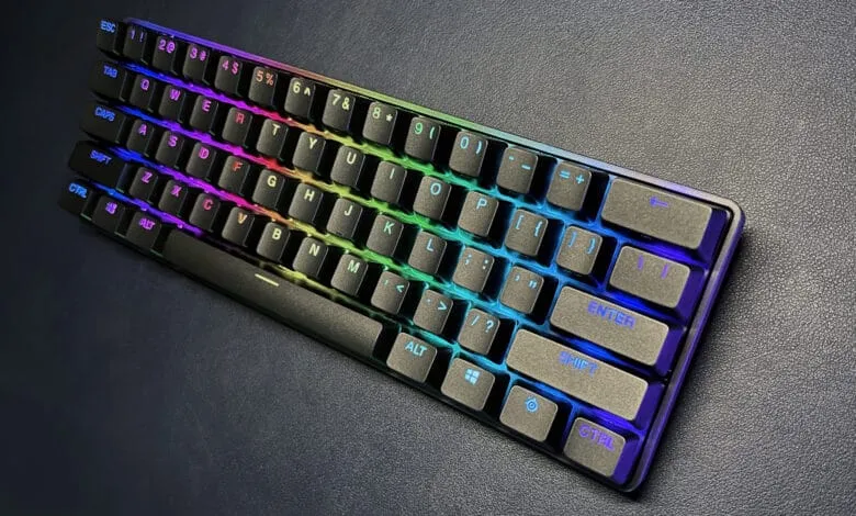 A compact, dark gray mechanical keyboard with RGB backlighting displays a rainbow spectrum across its keys.