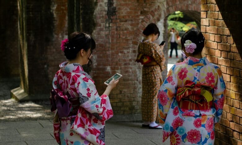 Three women in colorful kimonos use smartphones near a brick wall.