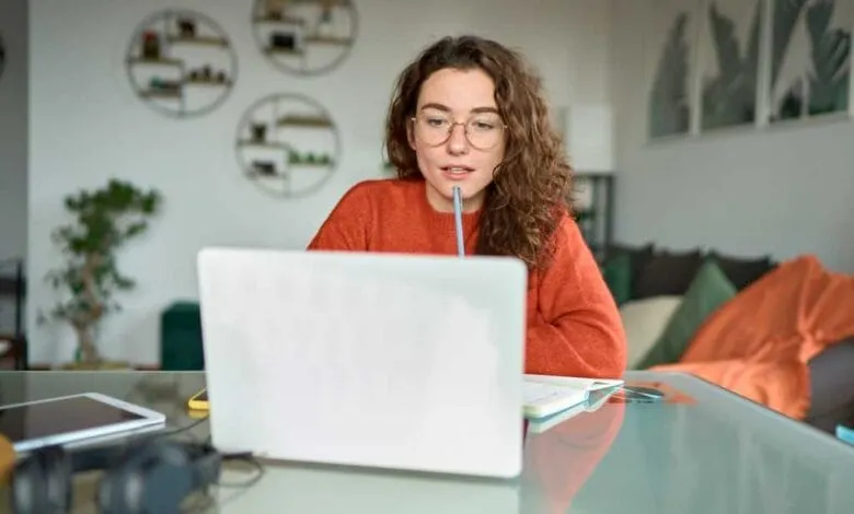 Young woman with curly hair and glasses works on her laptop at a glass desk in her home office.